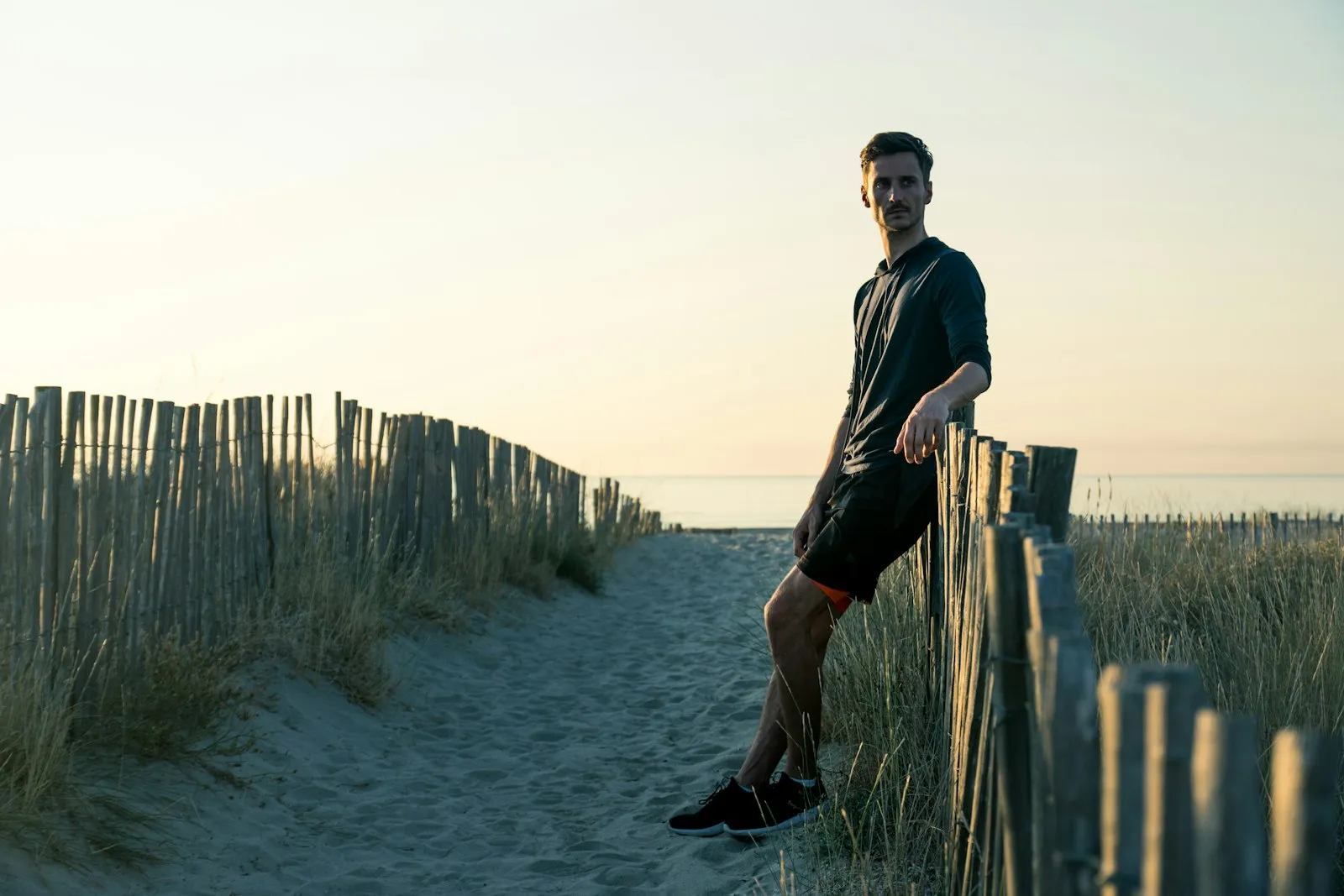A man in running clothes leaning on a wooden dune fence at golden hour, the sea behind him