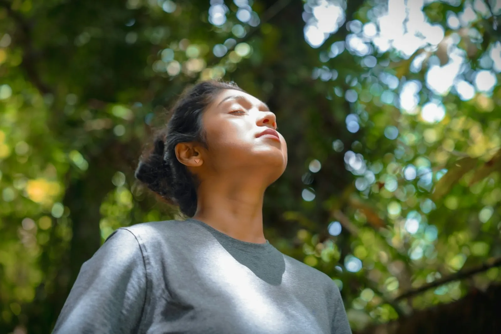 A woman with her eyes closed taking a slow breath under green trees, light falling on her face
