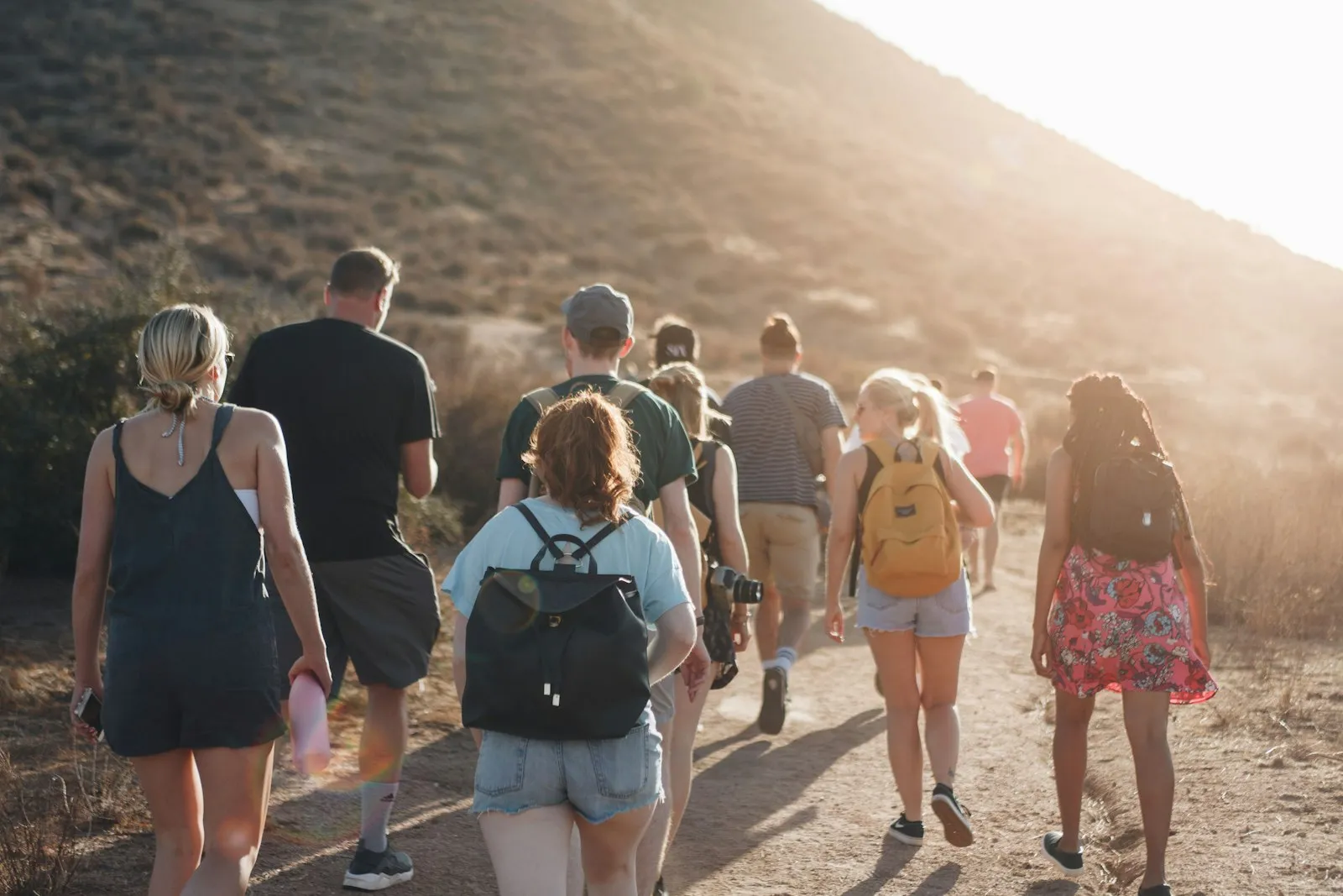 A group of friends seen from behind walking up a dirt trail on a sunlit hillside