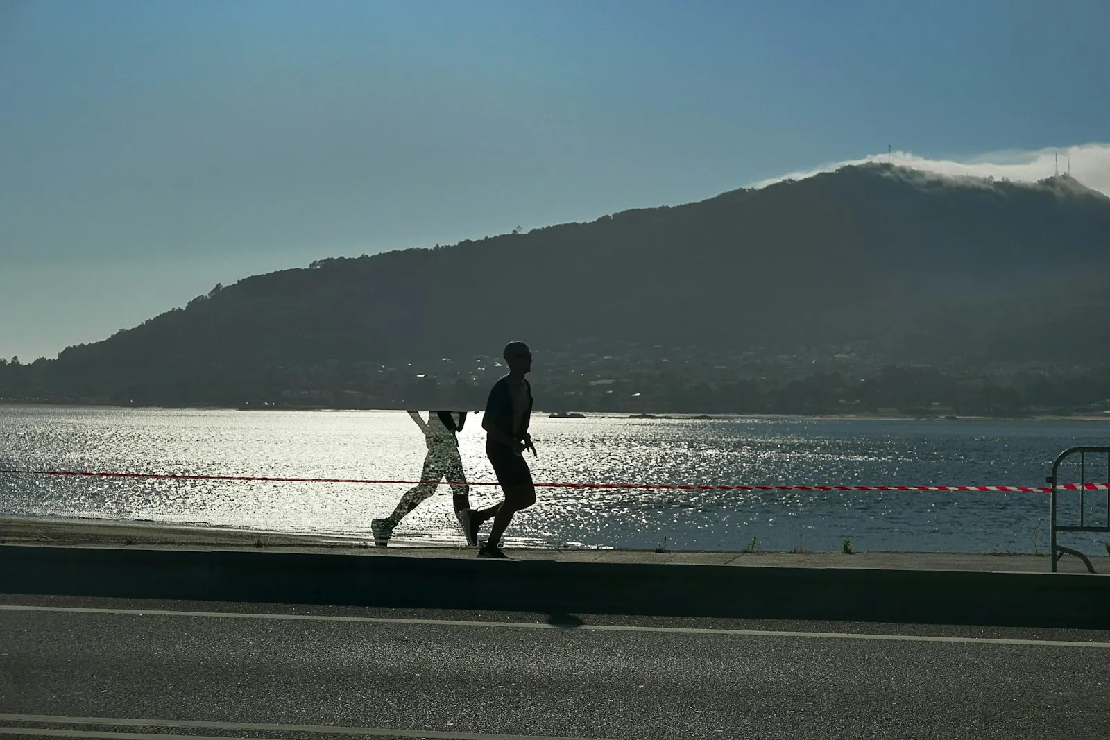 Two runners in silhouette on a lakeside road, light glittering on the water and a hill behind