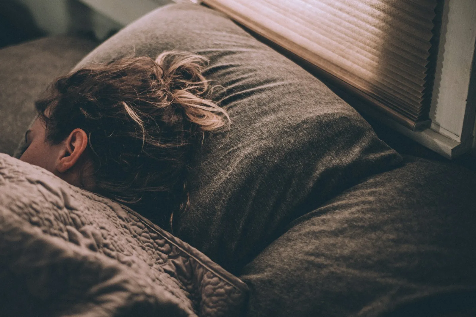 A person asleep face down in grey bedding, warm light coming through the blinds above