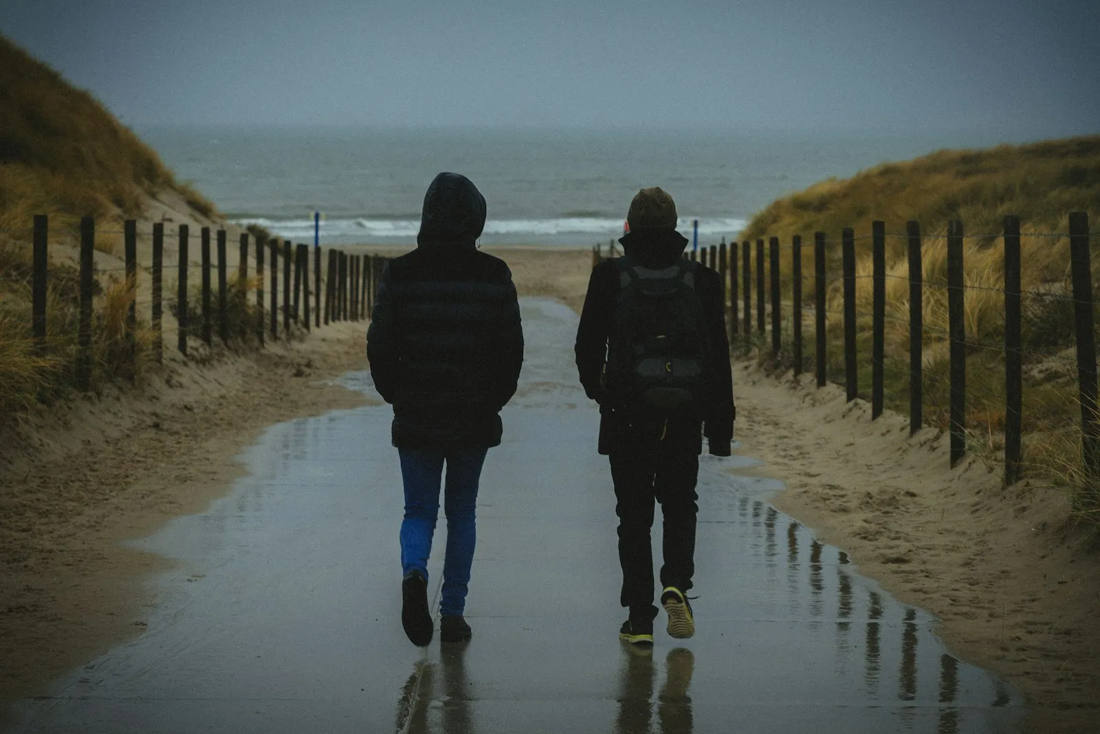 Two people in hooded jackets walking down a wet path between dunes toward a grey sea